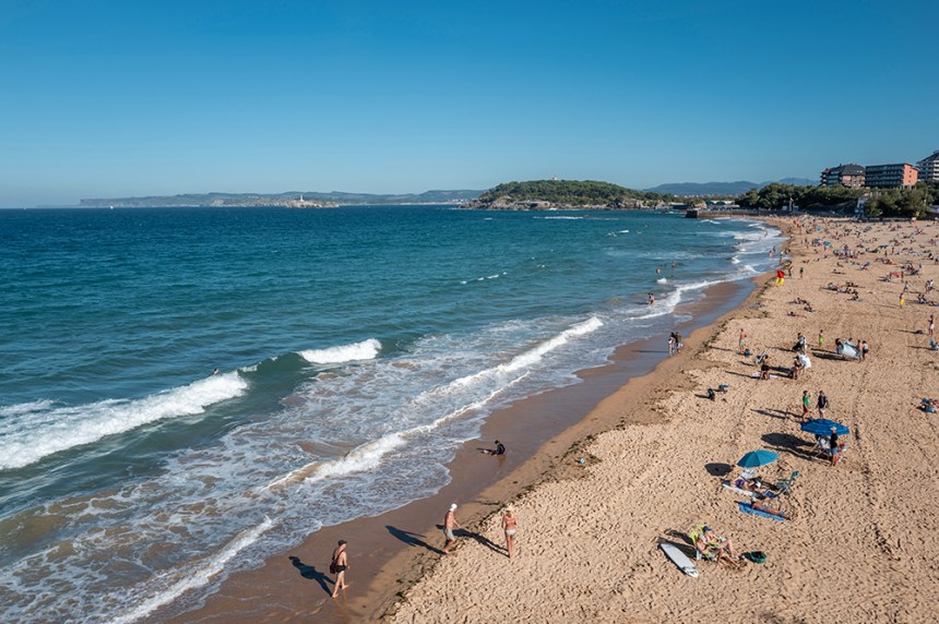 Плаж с многобройни хора, слънце и вълни, ясна синя вода и пясък.